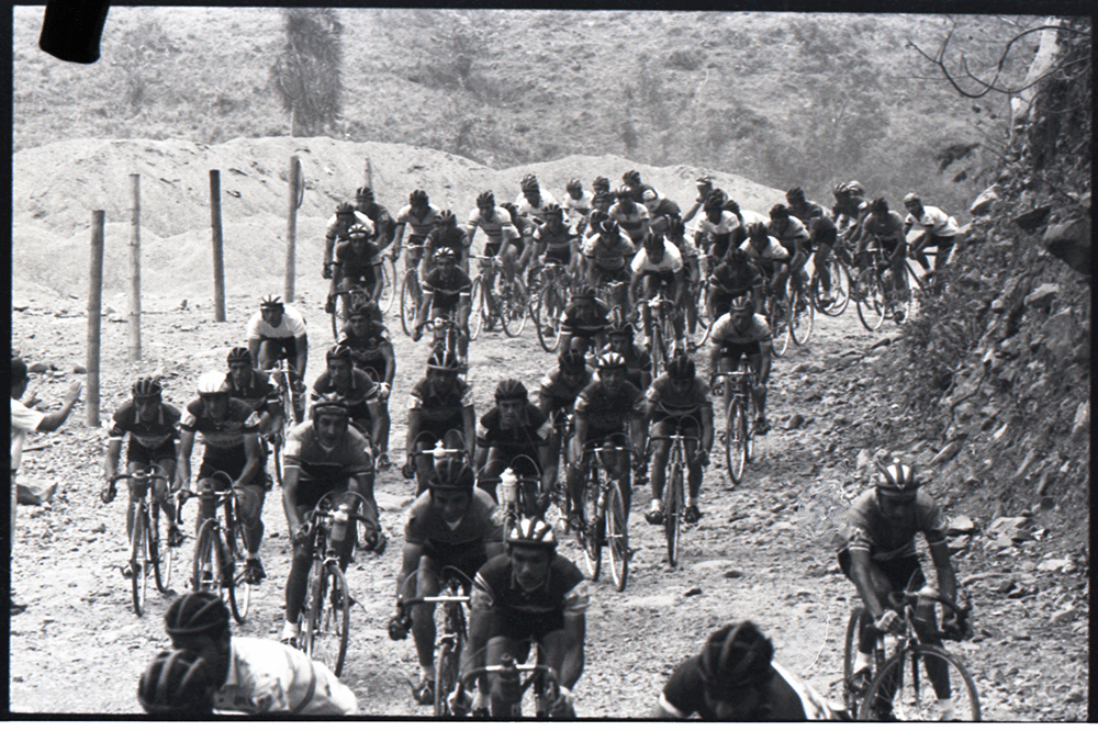 Vuelta a Colombia, s.f. Ciclistas en terreno destapado. Tal vez la expresión un 'río de ciclistas' nunca sea más apropiada que en el caso de esta imagen. La vuelta a Colombia tuvo un valor fundamental en la creación de una conciencia nacional del territorio. Esta imagen nos muestra las muy difíciles condiciones que encontraron los primeros competidores de las vueltas a Colombia, territorios aún sin vías pavimentadas por las que estos ciclistas forjaron la práctica de un deporte que ha devenido símbolo nacional.