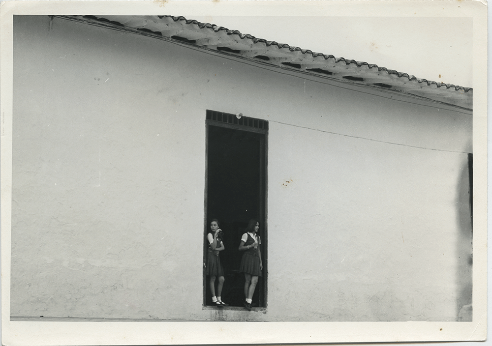 Niñas en el marco de una casa. Una fotografía tomada desde la distancia que nos muestra a dos niñas, ya acaso adolescentes, en lo que podría ser la puerta de su casa o una tienda, o un colegio. Suspendidas entre el adentro y el afuera, entre lo privado y lo público, entre la niñez y la juventud, en un instante casual y desprevenido que paradójicamente pareciera capturar algo esencial de ellas.