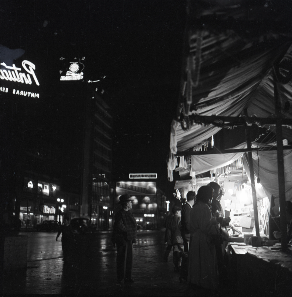 En esta fotografía, Caicedo captura una escena de un mercado nocturno, ubicado probablemente en la carrera 7 durante en un diciembre bogotano. La decoración con girnaldas del mercado da pistas sobre la época del año (ca. 1955). La escena juega con el contraste de luces producido por la oscuridad de la noche en contraposición a la luz artificial de la publicidad y las luces de los puestos del mercado. 