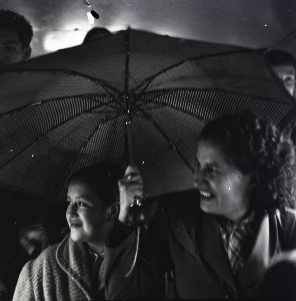 En esta escena Caicedo captura a dos mujeres mirando algo por fuera de la composición de la fotografía mientas sostienen un paraguas que las protegene de la lluvia. En la parte posterior se ven los rostros de dos hombres jóvenes lo que nos indica que quizá están en medio de mucha más gente. Caicedo juega con la composición de la fotografía dando protagonismo a las mujeres en el primer plano y aprovecha los constrastes de luz que produce la noche para iluminar la escena. 