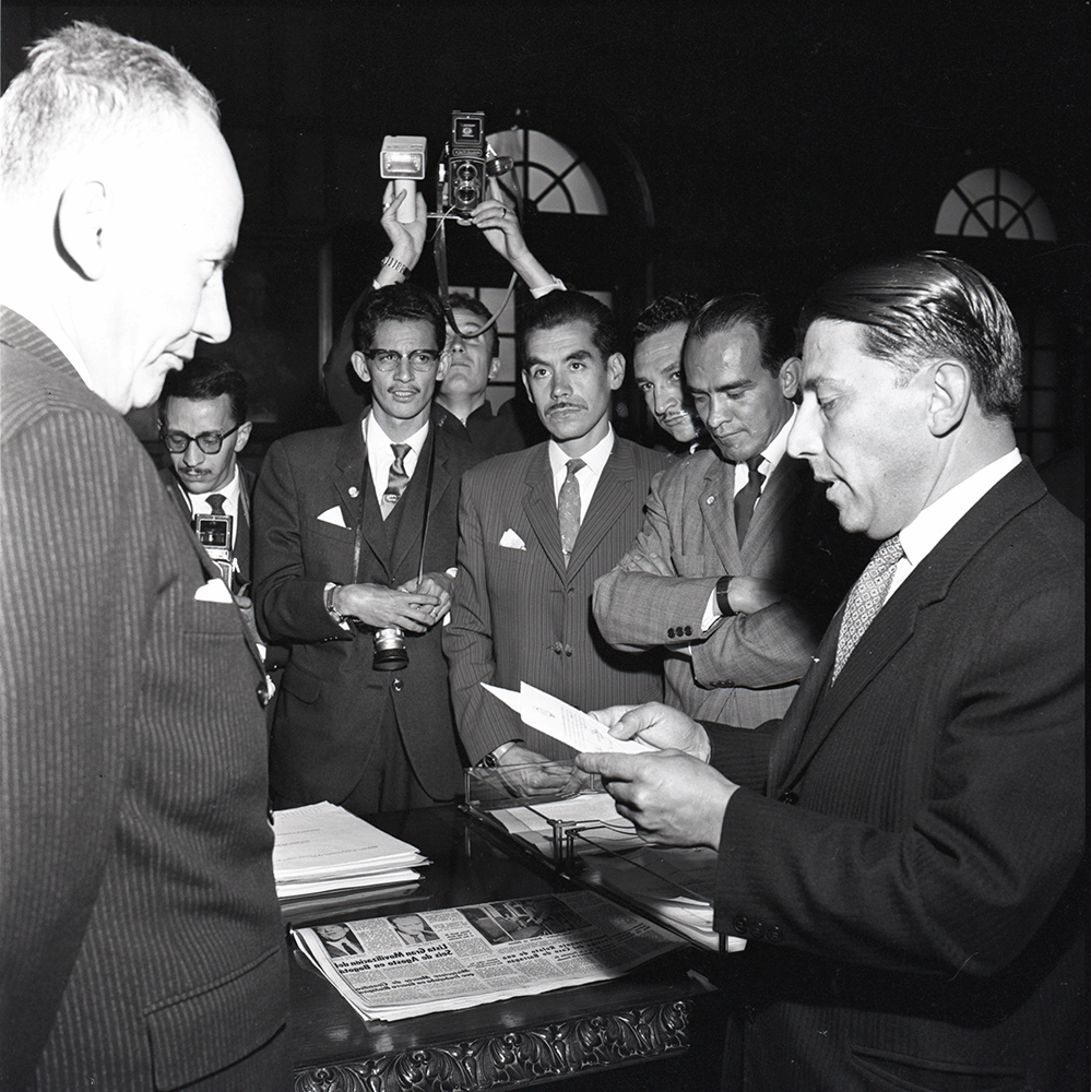 Alberto Lleras Camargo (1906-1990) fue el primer presidente del llamado 'Frente Nacional en Colombia, habiendo sido uno de sus proponentes, junto con Laureano Gómez. Años antes, por renuncia de Alfonso López Pumarejo a la presidencia, Lleras Camargo ejerció como presidente designado entre 1945 y 1946. Luego, fue presidente, elegido por elección popular, entre 1958 y 1962, primer periodo del Frente Nacional. Fue liberal y ejerció como periodista, columnista y como representante de Colombia ante la OEA. Fue figura clave en la relación entre Estados Unidos y Colombia, habiendo recibido en su presidencia préstamos de Estados Unidos para empezar un programa agrario que luego se convertiría en la Reforma Agraria del Frente Nacional. Bajo su segundo mandato se llevó a cabo la Alianza para el Progreso en Colombia. En la foto, vemos a Lleras Camargo como presidente en algún acto con reporteros gráficos. Circa 1959. Posiblemente reunidos en la Casa de Nariño. Información basada en el texto de Leopoldo Villar Borda, Alberto Lleras. El último republicano, Bogotá: Editorial Planeta, 1997. Sobre los préstamos, ver “Nuevos créditos a Colombia por 50 millones de dólares,” en El Tiempo, April 7, 1960. Archivo de Alberto Lleras Camargo, Biblioteca Luis Ángel Arango, 'El primer gobierno del Frente Nacional, Tomo III, Junio 13/59-April 9/60'.